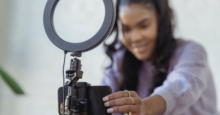 Tutorial Videos - Cheerful young African American female blogger in stylish sweater smiling while setting up camera of smartphone attached to tripod with ring light before recording vlog